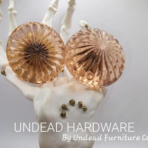 May include: Two round, copper-colored cabinet knobs with a ribbed design are displayed. The knobs are held by a white, skeletal hand. The text "UNDEAD HARDWARE By Undead Furniture Co" is at the bottom.