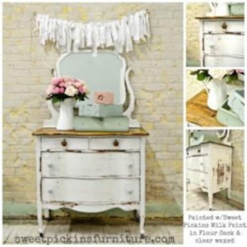 May include: A white antique dresser with four drawers and a distressed finish. The dresser has a wooden top and a mirror with a curved top. The dresser is decorated with a white ribbon garland and a vase of pink flowers.