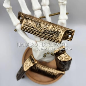 May include: Ornate, antique-style brass drawer pulls and handles with a floral design. The hardware is displayed on a white skeleton hand and a wooden tray. The text "UNDEAD HARDWARE By UNDEAD FURNITURE Co" is visible.