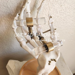 May include: A pair of clear glass cabinet knobs with brass hardware mounted on a white skeletal hand. The hand is on a wooden base. The text "UNDEAD HARDWARE by Undead Furniture Co" is visible on the base.