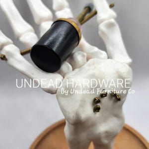 May include: A black and gold skeleton hand holding a black knob with a gold screw. The skeleton is standing on a wooden base. The text "UNDEAD HARDWARE By Undead Furniture Co" is printed below the skeleton.