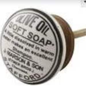 May include: A round, white and black knob with the text "OLIVE OIL SOFT SOAP" and "MARSON & SON" printed on it. The knob has a wooden base and a wooden stem. The knob is likely for a drawer or cabinet.