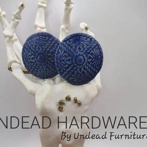 May include: Two blue ceramic earrings with a white and blue patterned design. The earrings are displayed on a white skeletal hand. The text "UNDEAD HARDWARE By Undead Furniture Co" is visible below the earrings.
