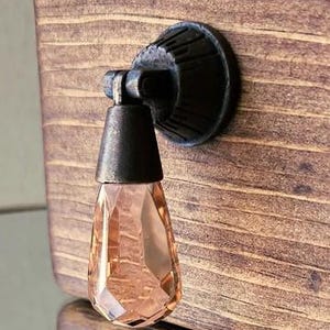 May include: A decorative cabinet pull with a faceted, teardrop-shaped, peach-colored crystal. The pull is attached to a dark, textured metal base and a dark metal stem. The pull is mounted on a wooden cabinet with visible wood grain.