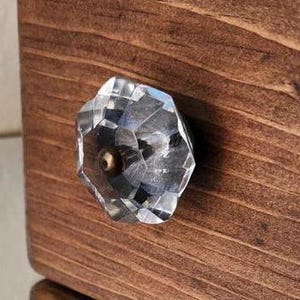 May include: A clear glass drawer knob with a faceted design and a brass screw. The knob is mounted on a dark brown wooden drawer.