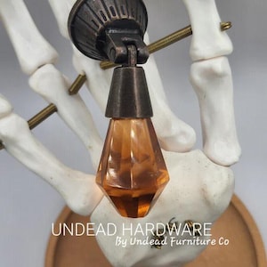 May include: A decorative lamp with a bronze finish and an amber-colored, faceted glass bulb. The lamp is mounted on a white skeleton hand. The text "UNDEAD HARDWARE By Undead Furniture Co" is visible.