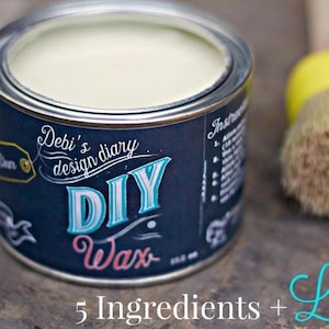 May include: A round metal can with a blue and white label that says "Debi's design diary DIY Wax". The can is filled with a white wax and is sitting on a wooden surface. There is a paintbrush with a yellow handle and brown bristles next to the can.