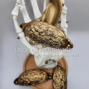 May include: Gold-colored cabinet hardware with ornate floral designs, displayed on a white skeleton hand. The image includes the text "Undead Hardware By Undead Furniture Co."