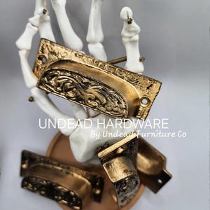 May include: Four gold-toned metal drawer pulls with an ornate design. The pulls are displayed on a white skeleton hand. The text "UNDEAD HARDWARE By Undead Furniture Co" is visible below the pulls.