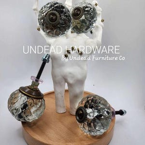 May include: Three clear glass cabinet knobs with a textured surface and brass accents. The knobs are attached to a white ceramic skeleton hand holding two of the knobs. The skeleton hand is standing on a wooden base. The text "UNDEAD HARDWARE by Undead Furniture Co." is printed below the skeleton hand.