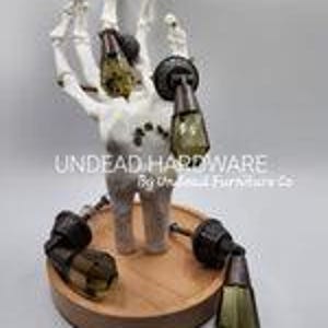 May include: Set of three skeleton hand shaped cabinet knobs with clear glass and brass accents. The knobs are displayed on a wooden base with the text "UNDEAD HARDWARE by The Deadwood Furniture Co."