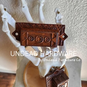 May include: Two wooden drawer pulls with intricate designs and brass accents. The pulls are displayed on a skeletal hand with the text "UNDEAD HARDWARE By Undead Furniture Co."