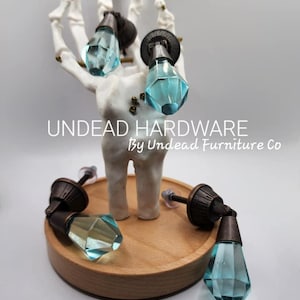 May include: Four antique-style cabinet knobs with a diamond-shaped glass insert. The glass is a light blue color. The knobs are displayed on a wooden base and a skeletal hand.