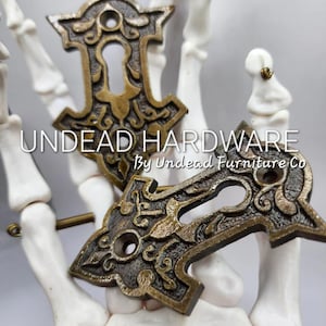 May include: Two ornate brass keyhole escutcheons with intricate floral designs. The escutcheons are displayed on a white skeletal hand. The text "UNDEAD HARDWARE By Undead Furniture Co." is visible below the escutcheons.