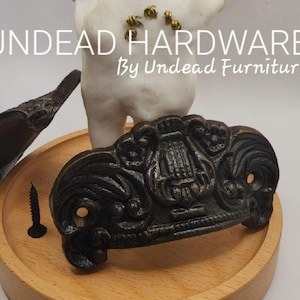May include: Two black metal drawer pulls with ornate designs. The larger pull has a lyre-shaped design in the center. The smaller pull is a simple curved shape. Both pulls are on a wooden surface. Undead Hardware by Undead Furniture Co.