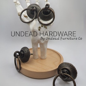 May include: Three antique bronze cabinet door pulls with ornate details. Each pull has a ring handle and a decorative plate. The pulls are displayed on a wooden surface and a skeletal hand.