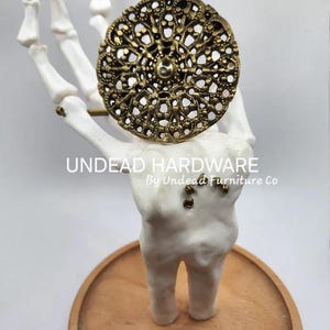 May include: A gold-toned metal ring with a decorative, intricate design. The ring is displayed on a white skeletal hand, which is standing on a wooden base. The text "UNDEAD HARDWARE by Undead Furniture Co." is printed below the hand.