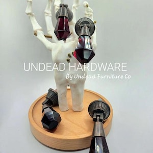May include: Four antique-style cabinet knobs with faceted glass teardrop-shaped handles. The knobs are displayed on a wooden stand and a skeletal hand. The text "UNDEAD HARDWARE By Undead Furniture Co" is visible below the knobs.