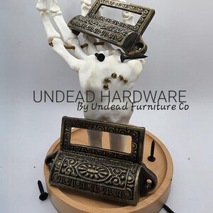 May include: Antique bronze-colored drawer pulls with ornate floral and scrollwork designs. The pulls are displayed on a wooden base with black screws, and the text "UNDEAD HARDWARE" is visible.