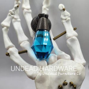 May include: A blue glass teardrop-shaped cabinet knob with a bronze metal base. The knob is held in a skeletal hand. The text "UNDEAD HARDWARE By Undead Furniture Co" is visible below the knob.