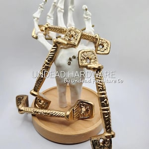 May include: Five gold-toned metal cabinet pulls with ornate designs. The pulls are arranged on a wooden base and a white skeletal hand.