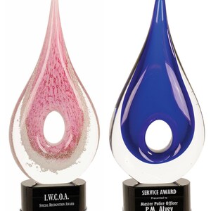 May include: Two glass teardrop awards with black bases. The award on the left is pink with white bubbles and the award on the right is blue. The award on the left has a black base with the text "I.W.C.O.A. Special Recognition Award". The award on the right has a black base with the text "SERVICE AWARD Presented to Master Police Officer P.M. Alvey".