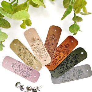 Peut inclure: Sept étiquettes en cuir avec un motif floral, dans différentes couleurs, notamment rose, marron, vert et gris. Les étiquettes ont un trou perforé en haut et sont disposées en éventail.
