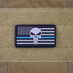 May include: A black and white American flag patch with a blue line and a white skull in the center. The patch is made of fabric and has a hook and loop backing.
