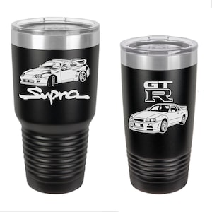 May include: Two black stainless steel tumblers with a white outline of a car and text. The tumbler on the left has a white outline of a Toyota Supra with the text "Supra". The tumbler on the right has a white outline of a Nissan Skyline GT-R with the text "GT-R".