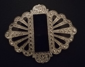 May include: A silver brooch with a black centre and a decorative design. The brooch has a fan-shaped design with a central rectangle.