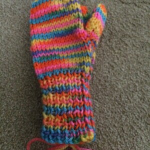 May include: A colourful striped knitted mitten with a pink, orange, yellow, blue and green colour scheme. The mitten has a thumb and is tied with a yellow and pink string.