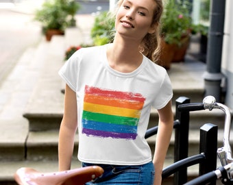 Può includere: Una maglietta bianca corta con un designo a bandiera arcobaleno. La bandiera è rovinata e ha un'estetica grunge. La maglietta è indossata da una persona con i capelli lunghi e biondi e jeans blu.