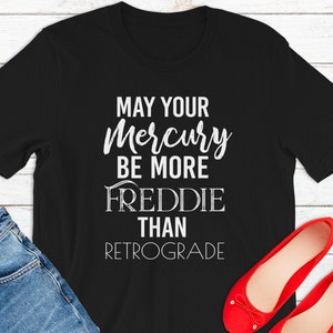 Può includere: Maglietta nera con testo bianco che recita "May your Mercury be more Freddie than retrograde". La maglietta è appoggiata su una superficie di legno con un paio di ballerine rosse e un paio di jeans blu.