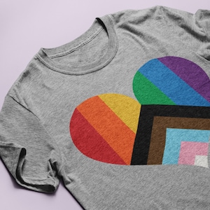Może przedstawiać: Szary T-shirt z wzorem tęczowego serca. Serce składa się z różnych kolorowych pasków, w tym czerwonego, pomarańczowego, żółtego, zielonego, niebieskiego, fioletowego, brązowego, jasnoniebieskiego, różowego i białego.
