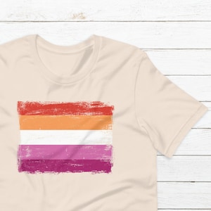 May include: A light beige t-shirt with a distressed lesbian pride flag design in red, orange, white, pink, and purple.