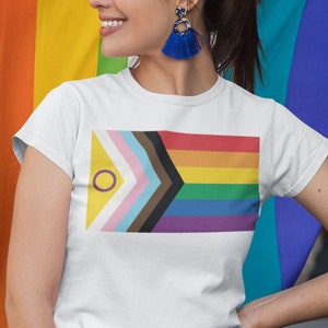 Puede incluir: Dos personas están de pie juntas sosteniendo una bandera del orgullo arcoíris. La persona de la derecha lleva una camiseta blanca con un diseño de bandera del orgullo y pantalones vaqueros azules. La persona de la izquierda lleva una camisa azul claro y pantalones cortos amarillos.