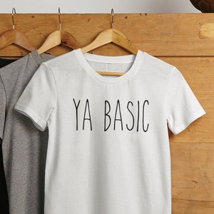 Op de afbeelding: Drie T-shirts hangen aan hangers. Het witte T-shirt heeft de tekst "YA BASIC" op de voorkant gedrukt. De andere twee T-shirts zijn zwart en grijs.