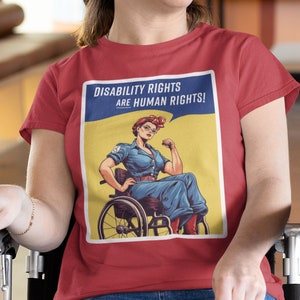 Könnte beinhalten: Eine Frau in einem roten T-Shirt mit einer Grafik im Vintage-Stil einer Frau im Rollstuhl mit dem Text "Disability Rights Are Human Rights!" Die Frau lächelt und schaut nach oben.