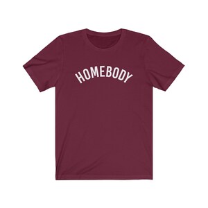 May include: A maroon t-shirt with the word "HOMEBODY" printed in white in an arc across the chest.
