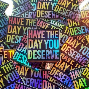 May include: A close-up of a pile of colorful stickers with the text "Have the day you deserve" printed on them. The stickers are iridescent and have a rainbow gradient effect.