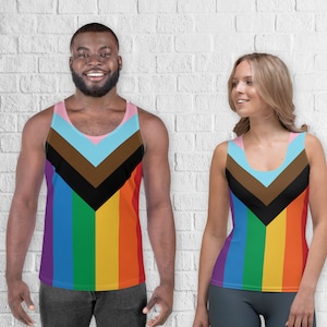 Puede incluir: Dos camisetas sin mangas con un diseño de arcoíris. Las camisetas presentan un patrón en forma de V en negro, marrón, azul claro y rosa, sobre rayas verticales de arcoíris. Las camisetas sin mangas las llevan dos personas, un hombre y una mujer.