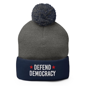 May include: A gray beanie with a navy blue brim and a dark gray pom-pom. The brim features the embroidered text "DEFEND DEMOCRACY" in white with red star accents. A winter hat with a political message.