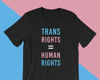 Könnte beinhalten: Schwarzes T-Shirt mit dem Text "TRANS RIGHTS = HUMAN RIGHTS" in Blau und Rosa. Der Text ist mittig auf der Vorderseite des Shirts platziert. Der Hintergrund ist in Rosa und Blau geteilt.
