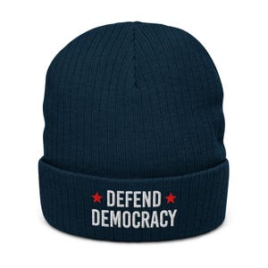 May include: A navy blue ribbed knit beanie with the words "DEFEND DEMOCRACY" embroidered in white, flanked by two red stars. The beanie is a classic winter accessory.