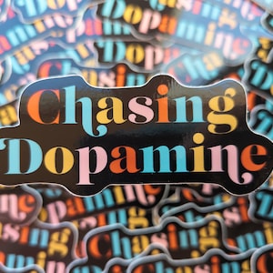 May include: A close-up of a sticker with the text "Chasing Dopamine" in a colorful, retro font. The sticker is black with yellow, orange, pink, and blue lettering. The sticker is surrounded by other stickers with the same design.