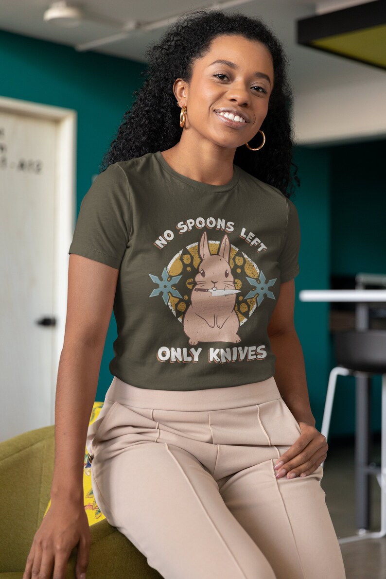 May include: A woman wearing a green t-shirt with a cartoon rabbit holding a knife and the text "NO SPOONS LEFT ONLY KNIVES".