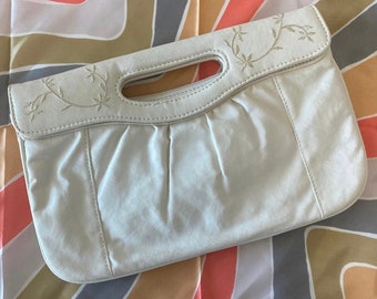 May include: Cream-colored clutch purse with a handle and floral embroidery. The purse has a pleated design and a rectangular shape. The handle is a cutout design. The purse is on a colorful patterned background.