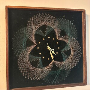 May include: A string art clock with a black velvet background. The clock face is surrounded by a design of pink and green string.
