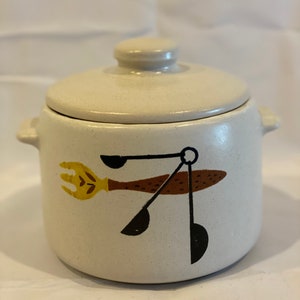 Peut inclure: Un pot en céramique blanche avec un couvercle, orné d'un motif d'ustensiles de cuisine en brun, jaune et noir. Le pot a un glaçage moucheté.