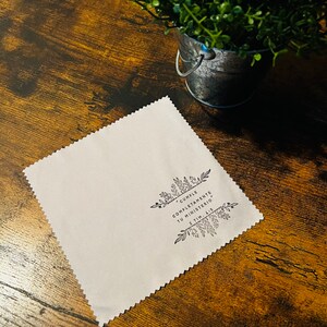 May include: A white microfiber cleaning cloth with a black floral design and the text "Cumple Completamente Tu Ministerio" 2 Tim. 4:5. The cloth is on a brown wooden surface.